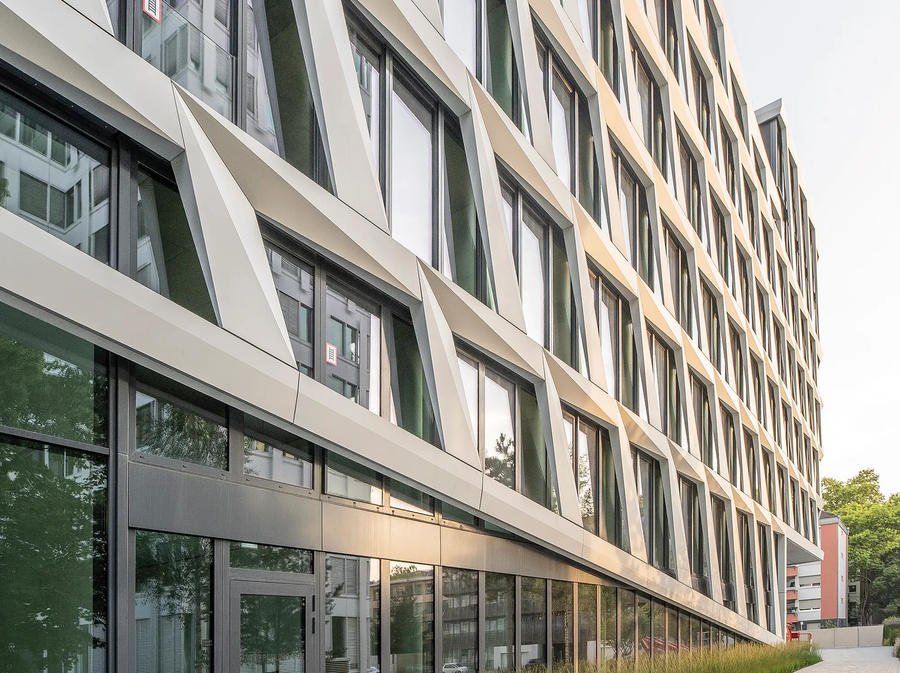 Seitenansicht des ONE PLAZA Gebäudes mit strukturierter Fassade, großen Fenstern und bepflanztem Vorbereich.